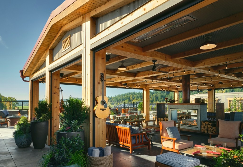 Covered rooftop terrace with timber beams, string lights, a fireplace, plants and lounge seating, looking out over green hills.